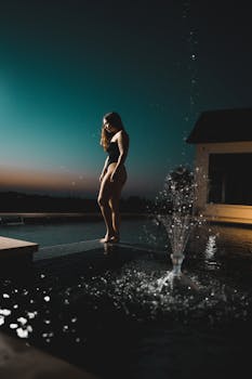 夜晚，一名身着泳衣的女子站在灯火通明的泳池边，捕捉着宁静的时刻