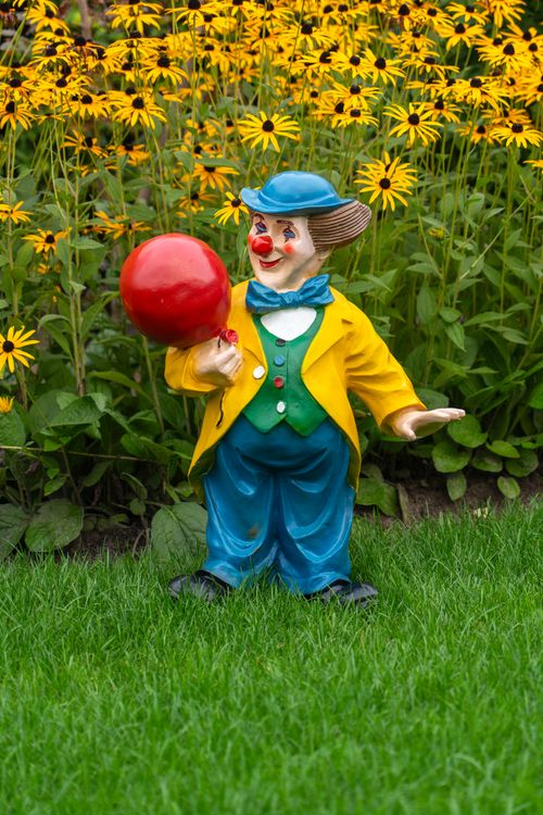 Free Colorful clown statue with red balloon among yellow flowers in a vibrant garden setting. Stock Photo