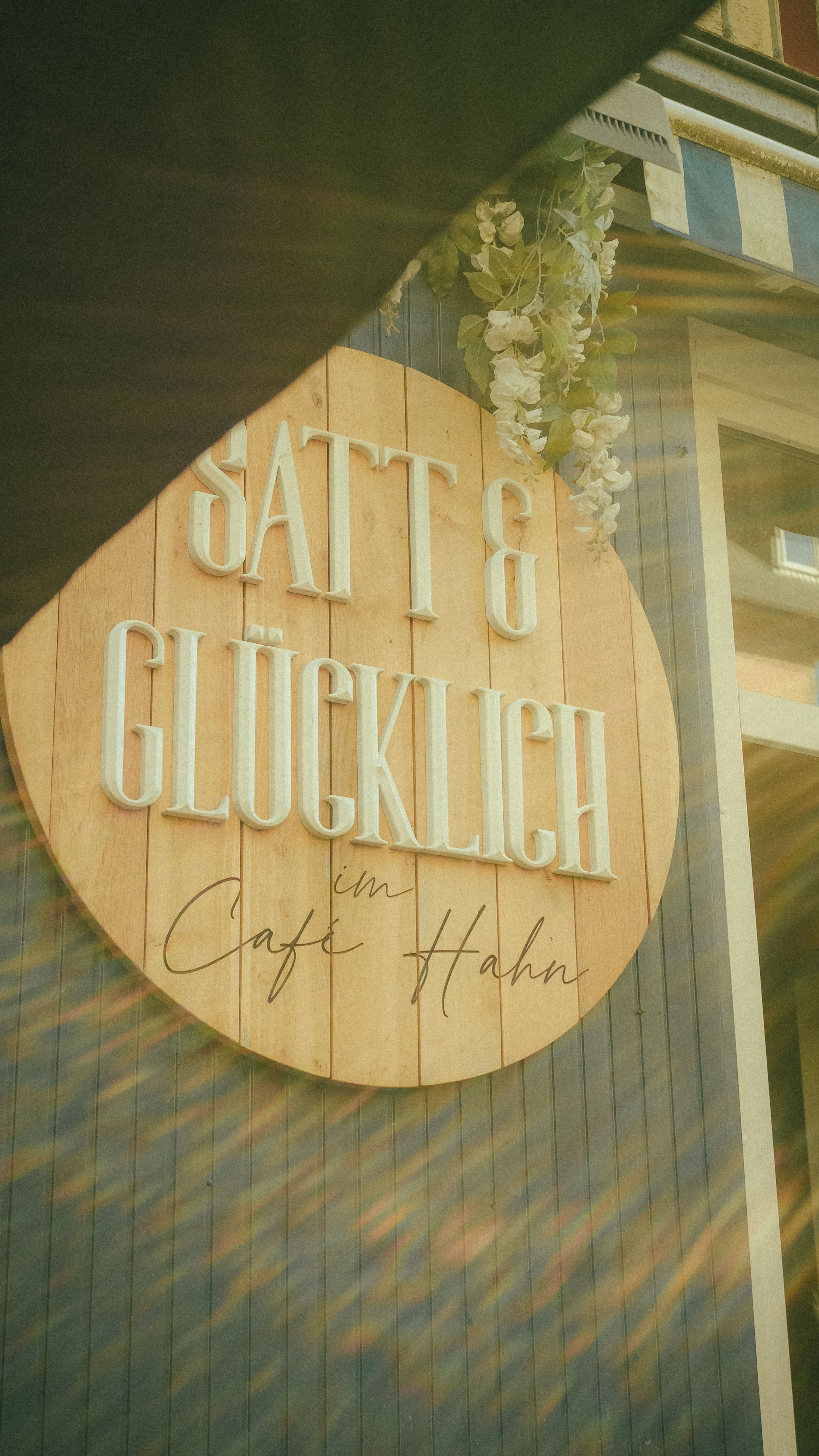 Charming cafe sign 'Satt & Glücklich' with sunrays and floral decor.