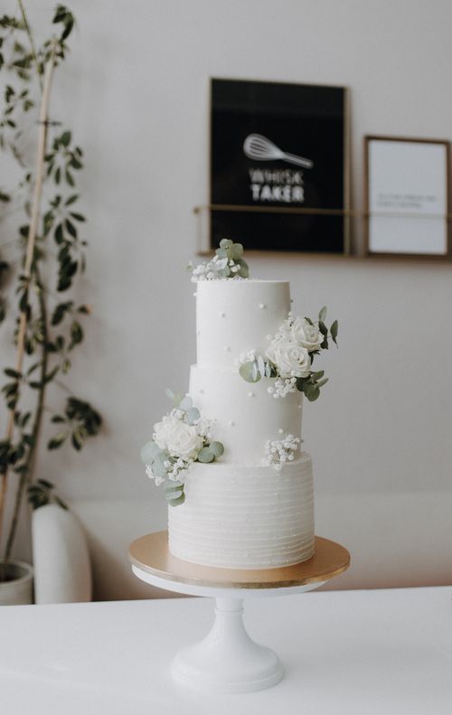 Free Elegant three-tier white cake adorned with white flowers and greenery, perfect for weddings. Stock Photo