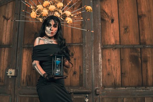 A stylish woman in Catrina makeup and costume holding an hourglass, marking Dia de los Muertos.