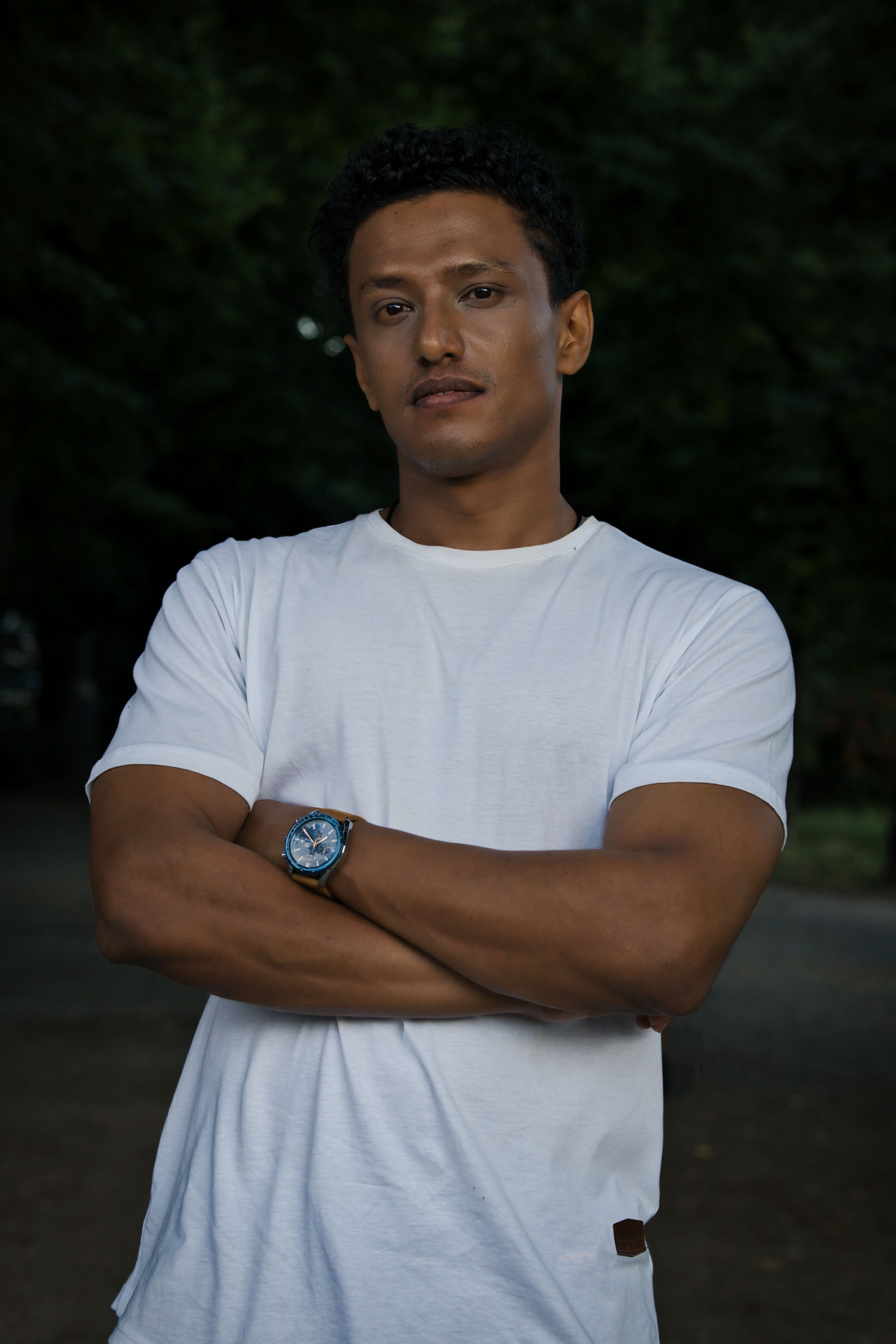 A confident young man crosses his arms and poses outdoors in Berlin, wearing a casual white tee.