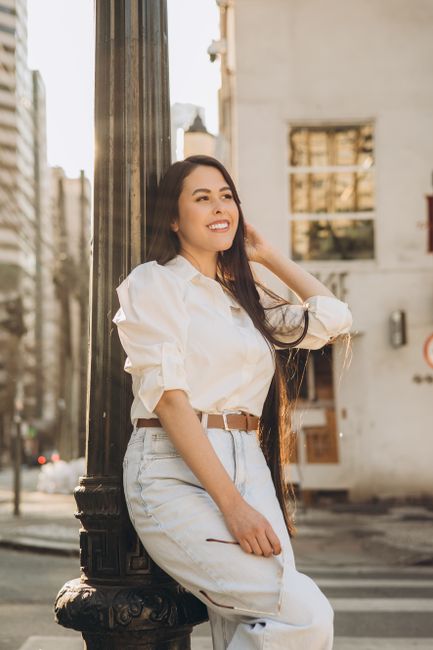 Casual portrait of a woman in white outfit leaning against a lamp post on a sunny city street.
