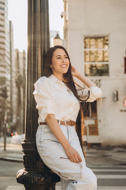 Free Casual portrait of a woman in white outfit leaning against a lamp post on a sunny city street. Stock Photo