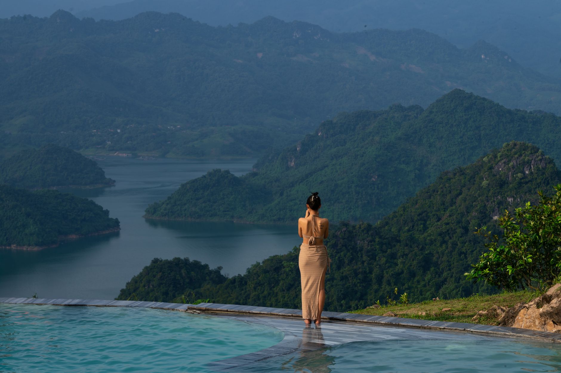 A serene view of Hoa Binh lake and mountains with a woman enjoying the infinity pool.