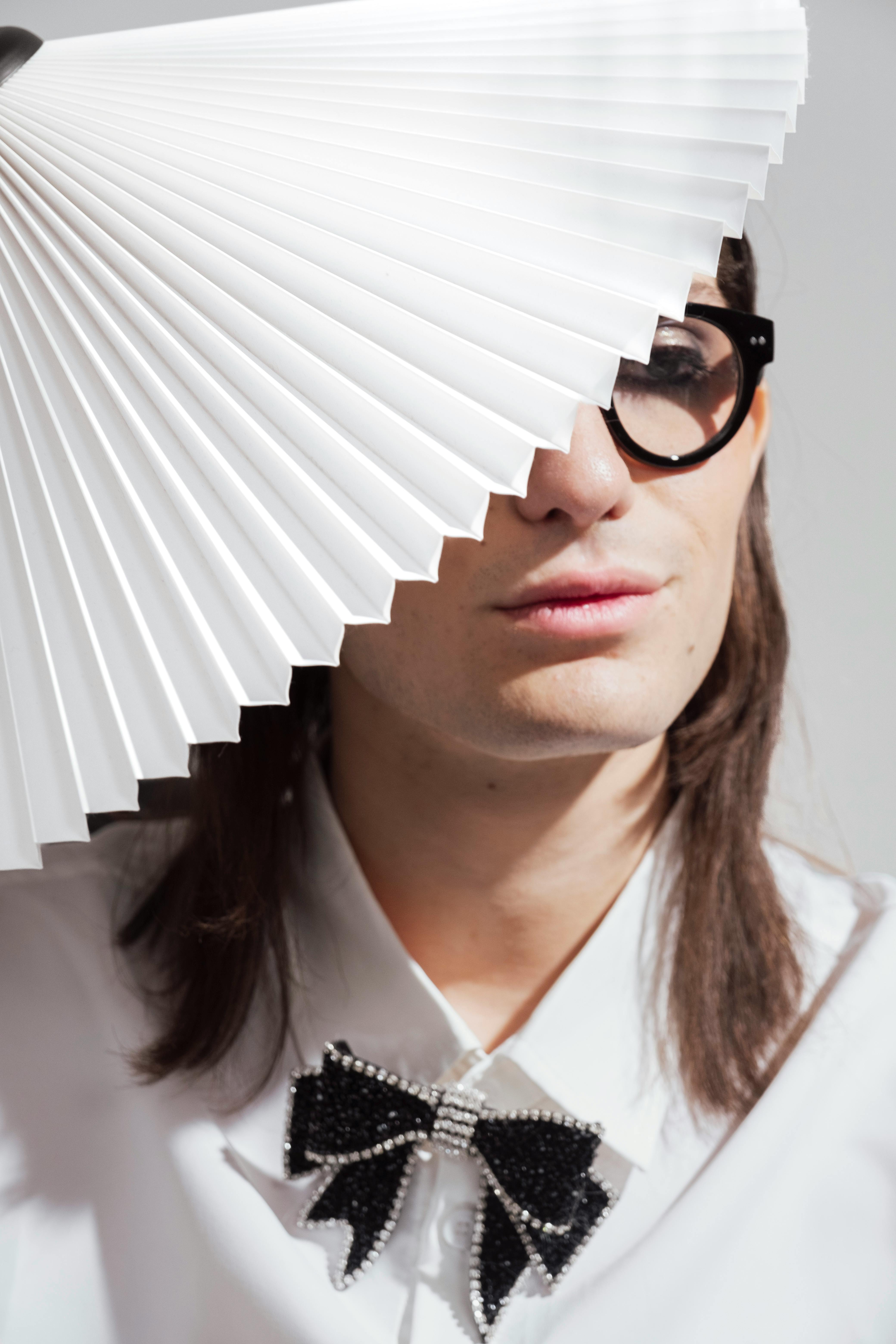 Stylish portrait of a person wearing large eyeglasses and a beaded bow tie, partially covered by a pleated fan.