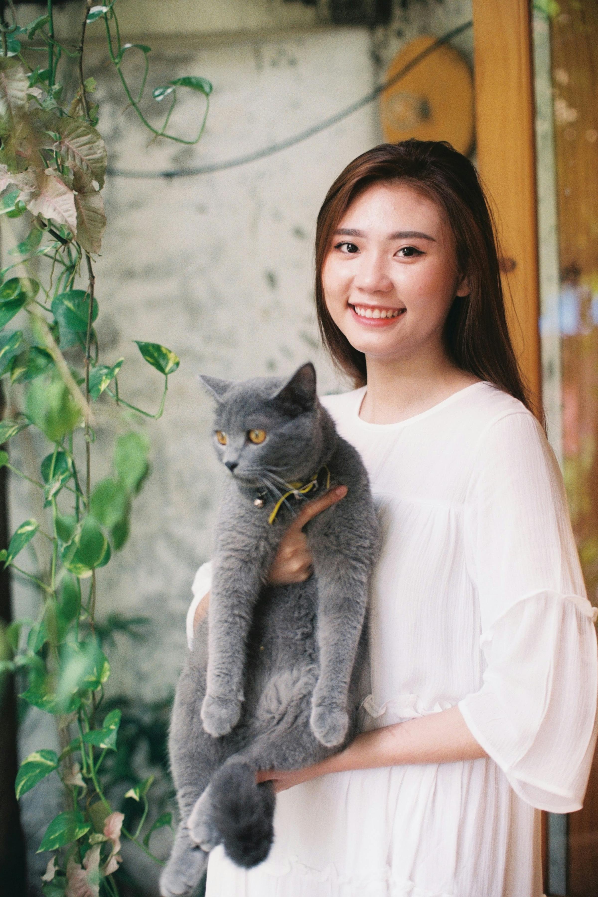 Smiling woman in white dress holding a grey cat indoors, surrounded by plants.