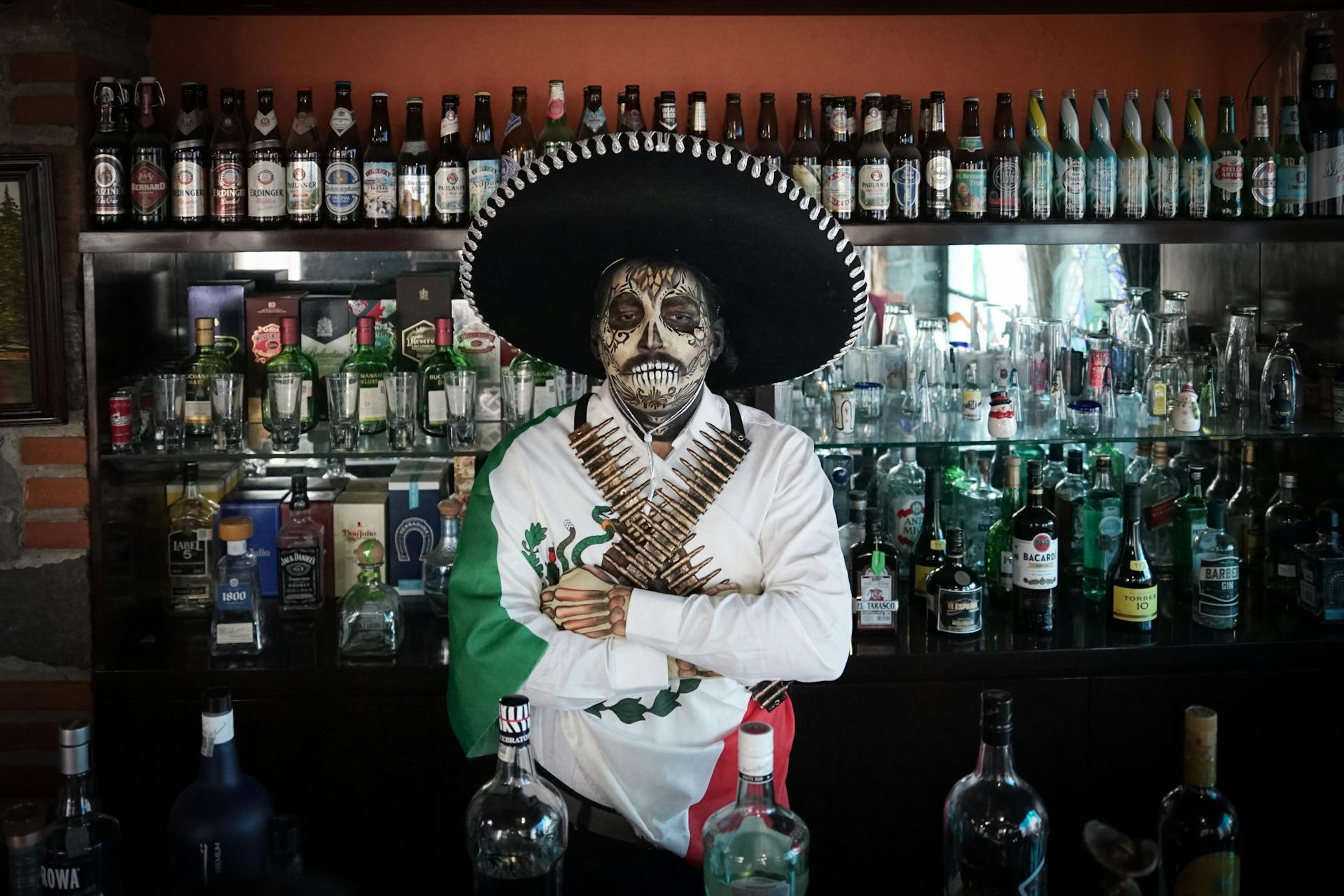 A bartender with a Catrina face paint and sombrero at a Mexican bar filled with spirits and decor.