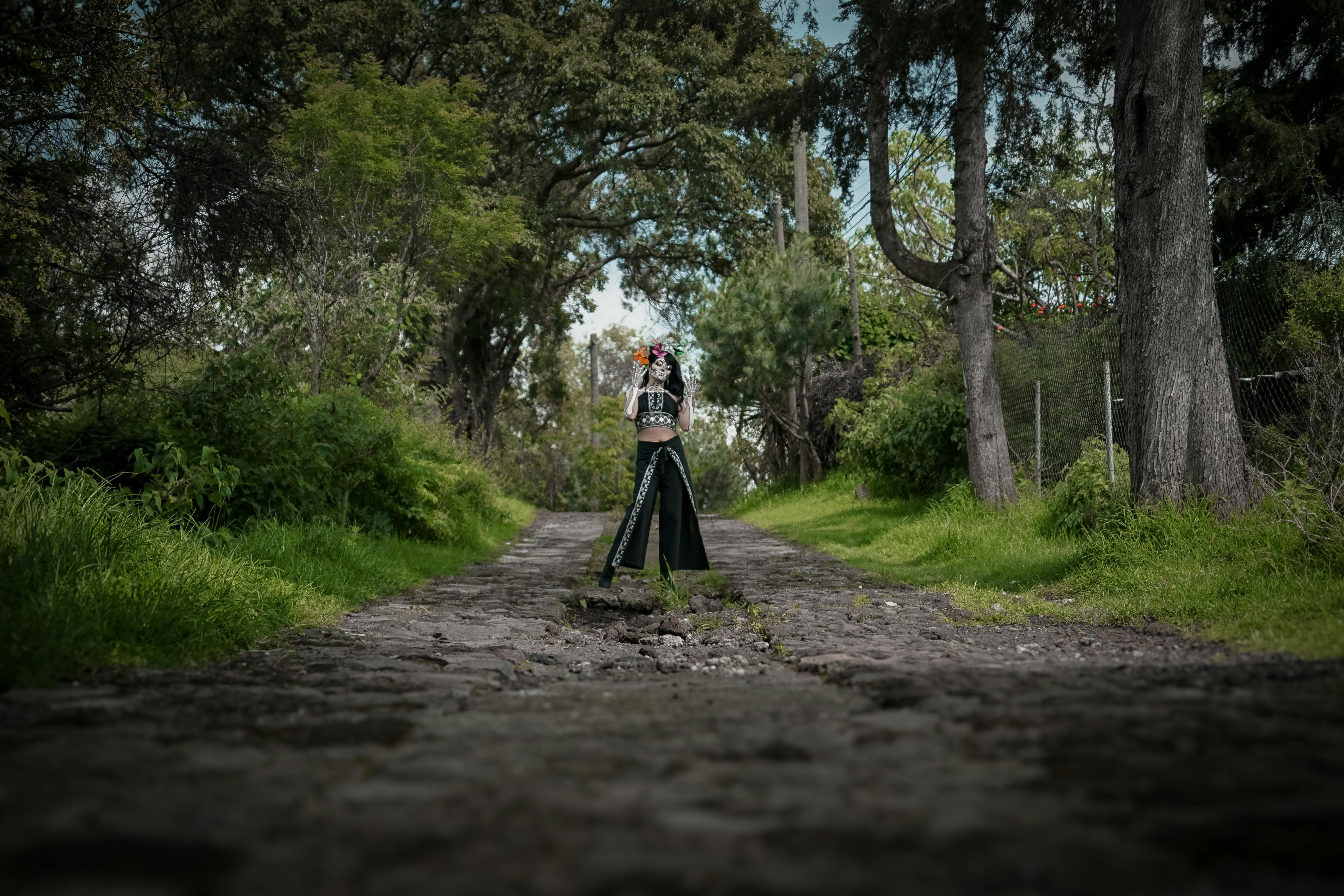 A lone woman in gothic attire walks down a tree-lined cobblestone path, surrounded by lush greenery.