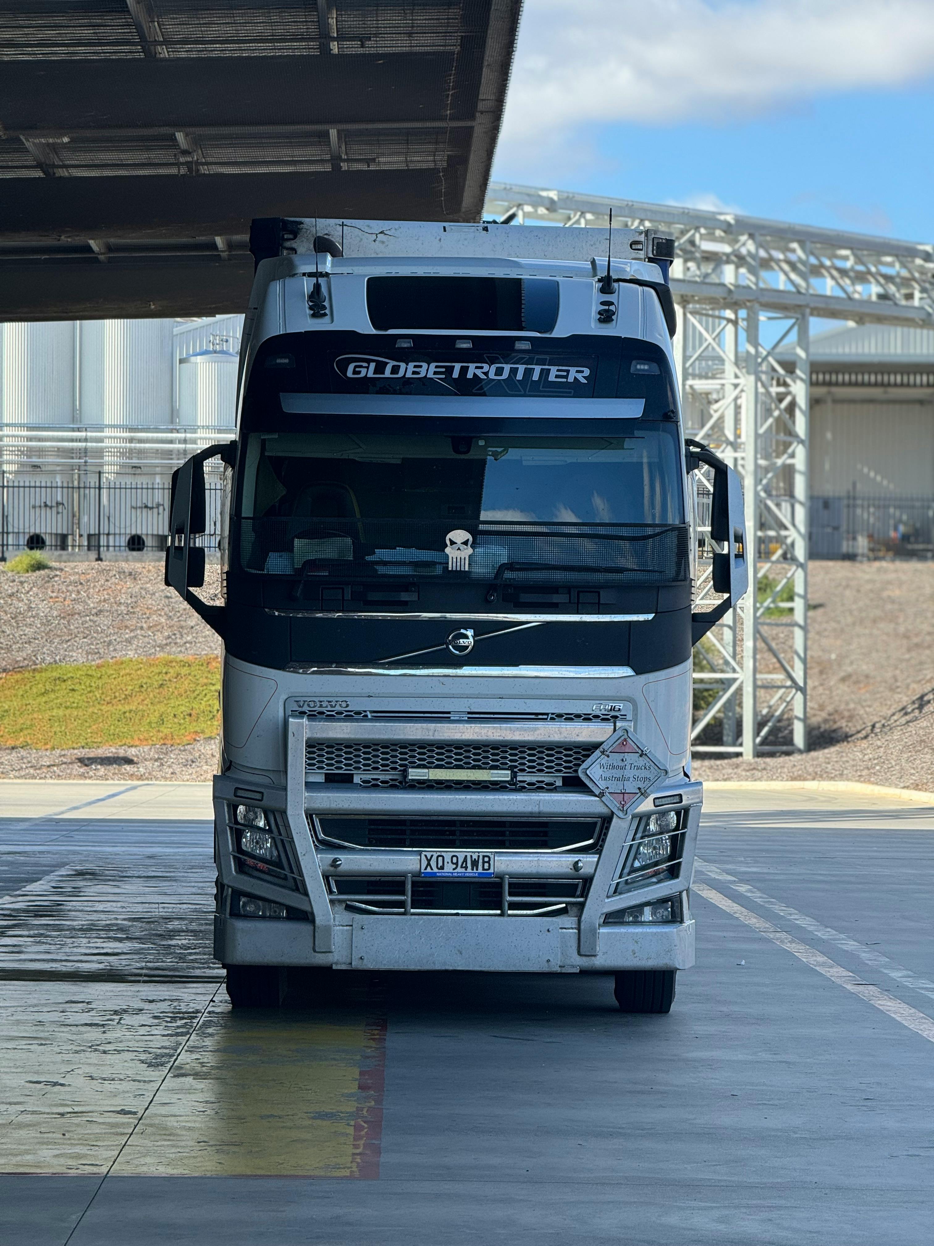 White semi-truck parked under a structure with a chrome front. “GLOBAL TRUCKER” is written on the visor.
