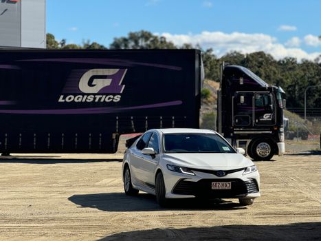 White sedan parked in front of a logistics truck at an outdoor loading area on a sunny day.