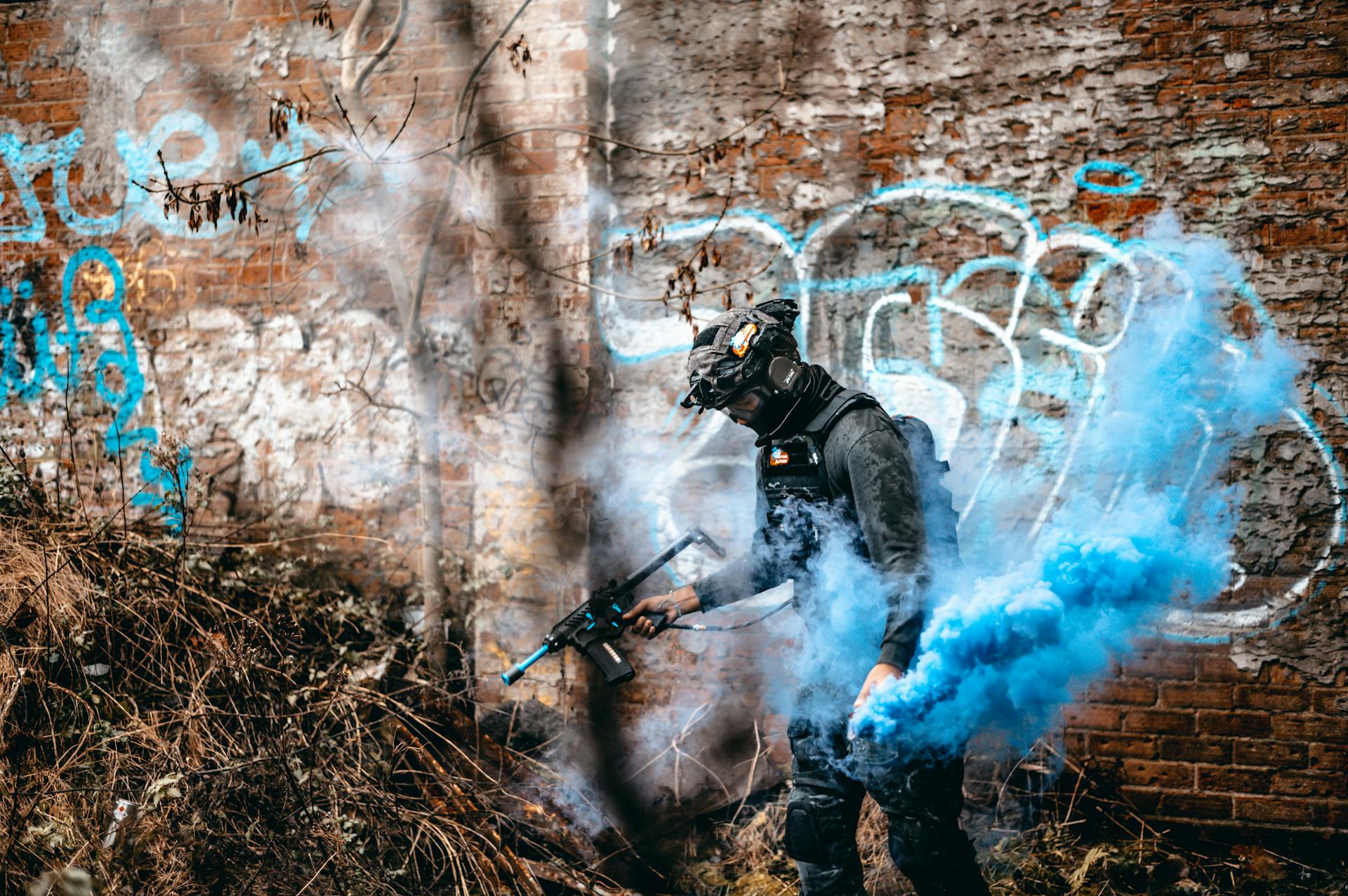 A person in tactical gear engages in an airsoft game with colorful smoke in an urban graffiti area.