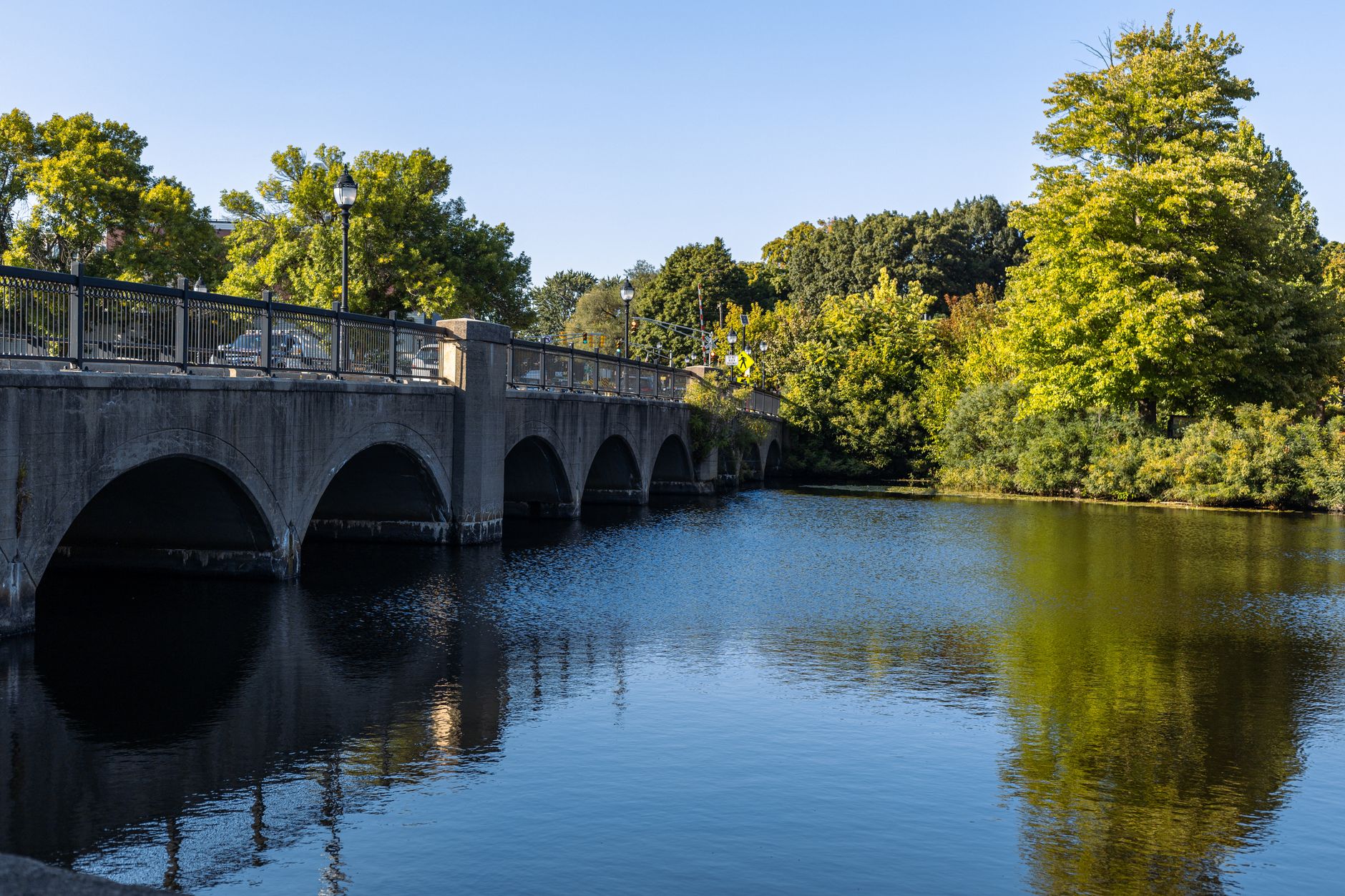 A picturesque stone bridge spans a calm river, reflecting lush green trees and a clear blue sky.