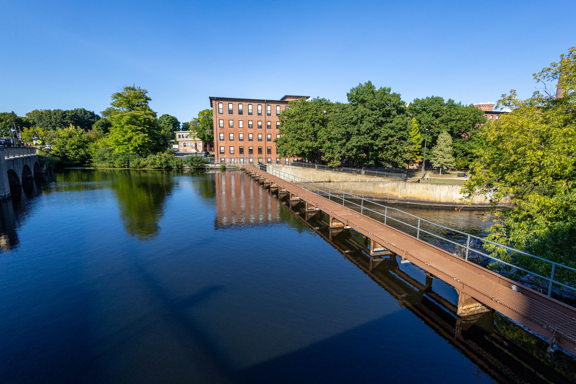 Historic building by a tranquil river with a bridge reflecting in the water, under clear blue skies.