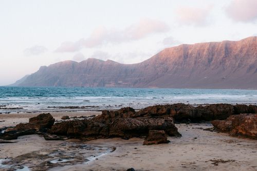Free Stunning coastal view featuring rocky shoreline, tranquil ocean, and majestic mountains at dawn light. Stock Photo