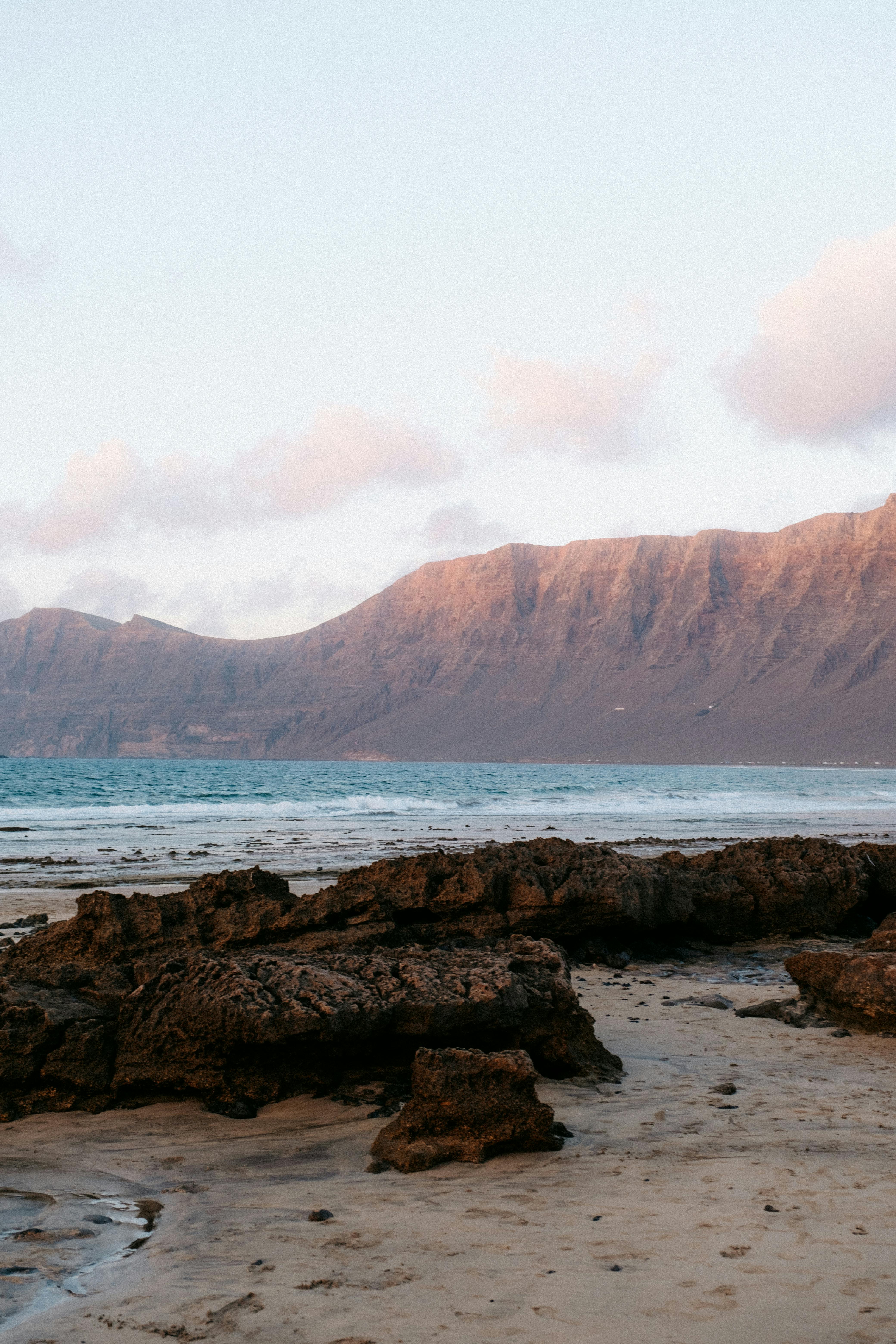 Tranquil beach scene at sunset featuring rocky shore and distant mountains under a pastel sky.