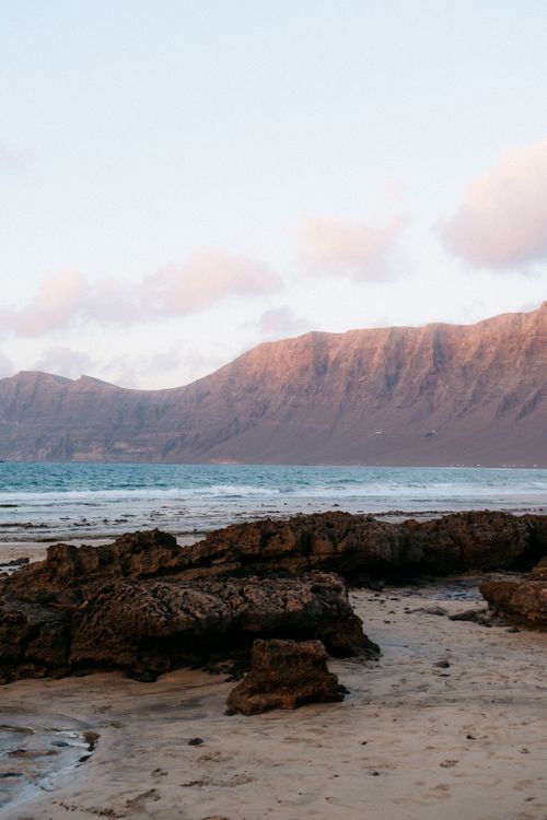 Free Tranquil beach scene at sunset featuring rocky shore and distant mountains under a pastel sky. Stock Photo