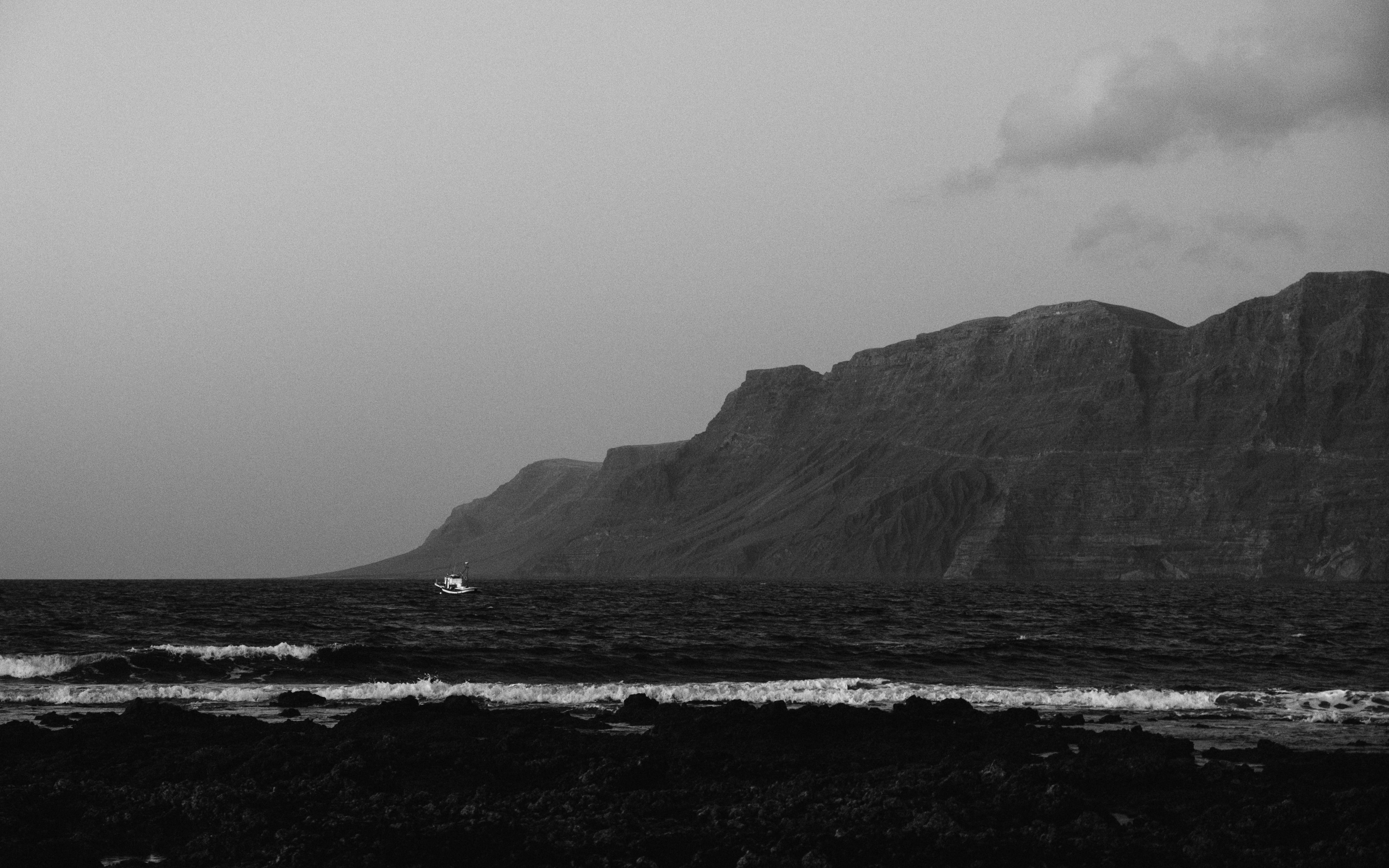A black and white view of rocky coast with waves and a distant boat.