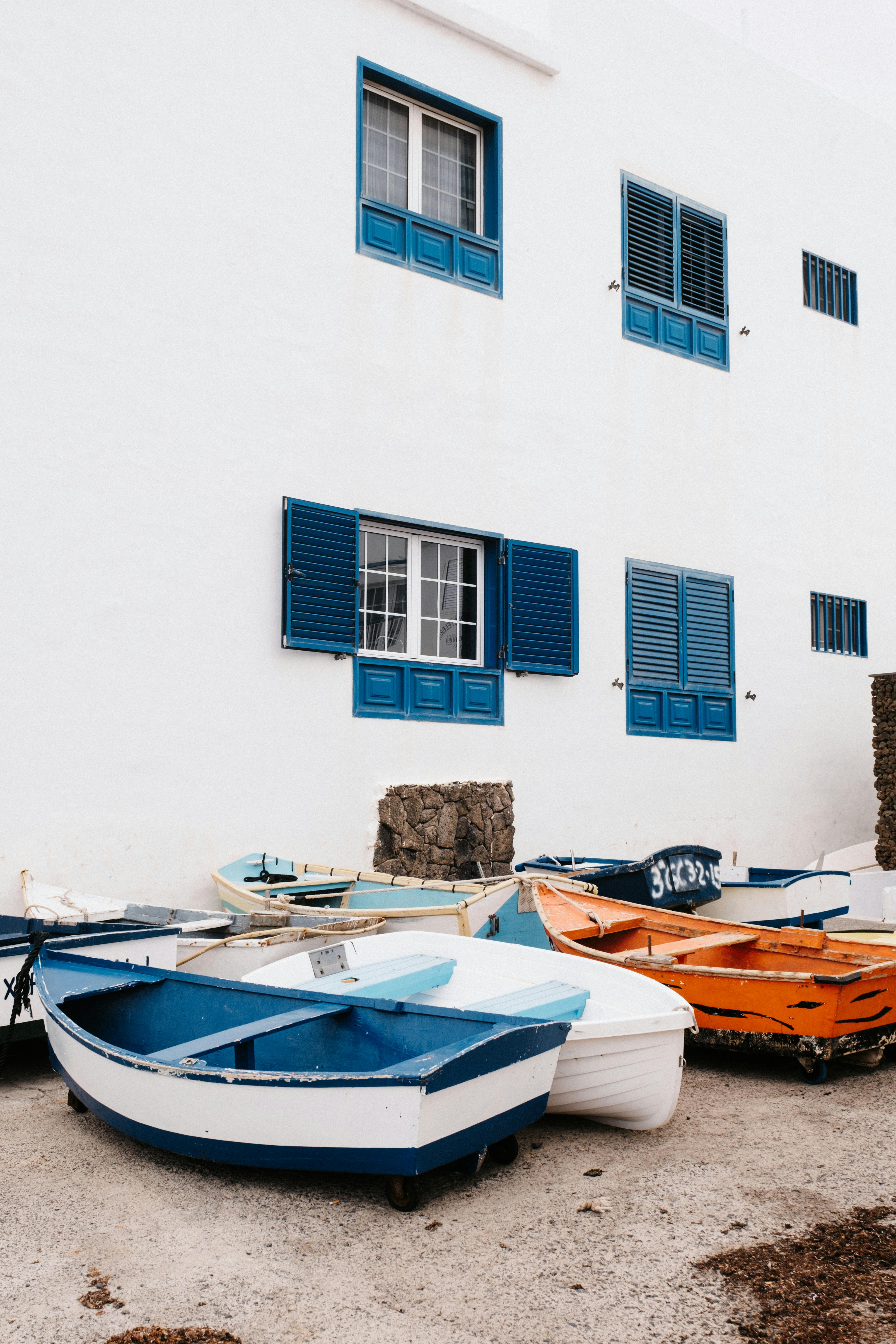 Rowboats resting against a white building with vibrant blue shutters and windows on a sunny day.