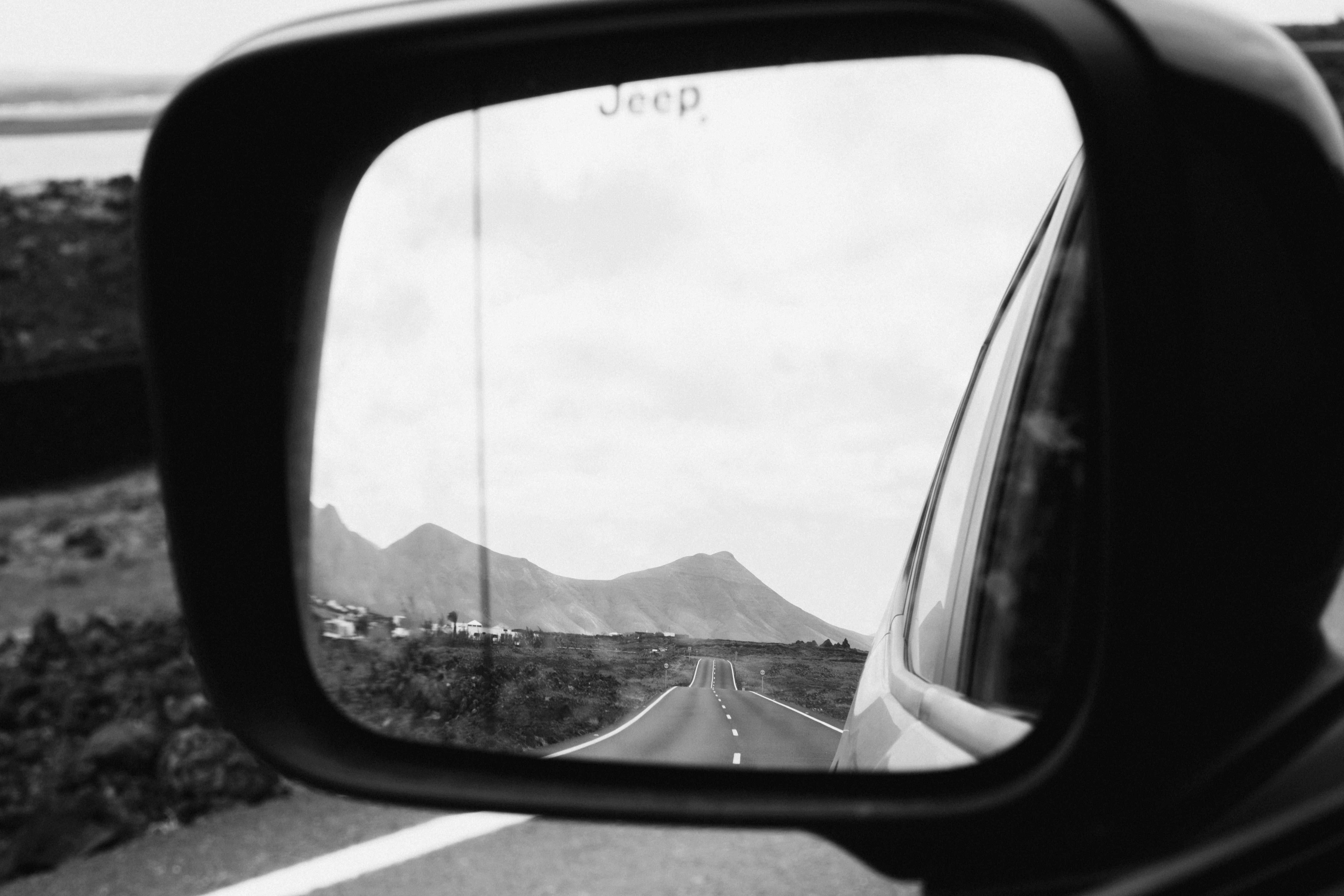 A black and white image capturing a scenic road through a car's rearview mirror.