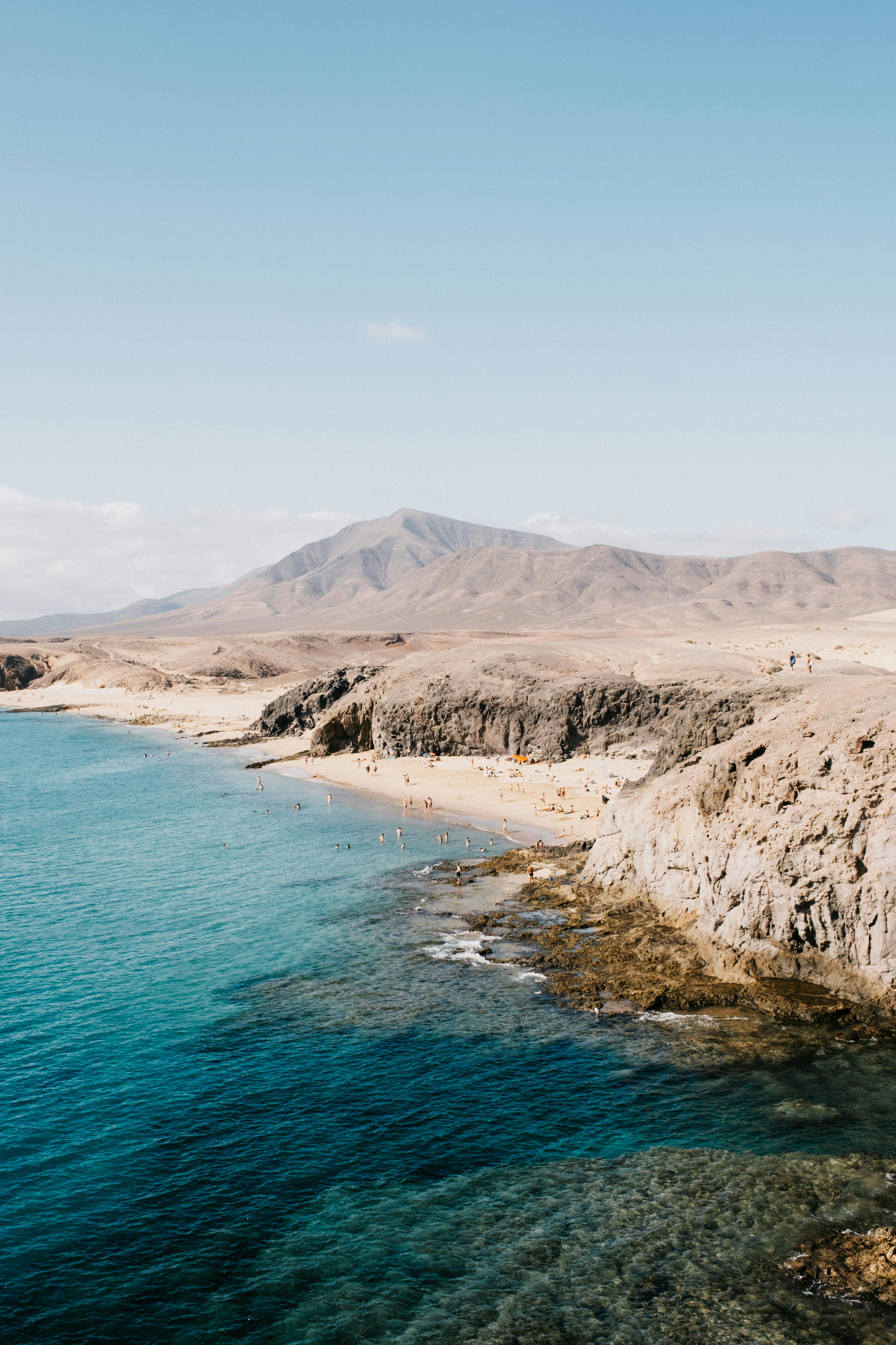 Stunning aerial view of a rocky coastline with clear blue ocean and distant mountains under a blue sky.