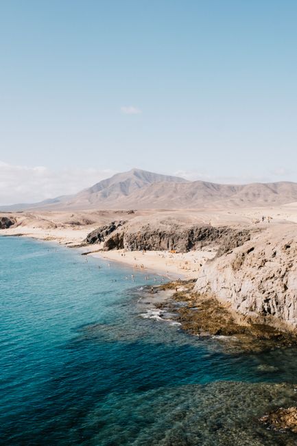 Stunning aerial view of a rocky coastline with clear blue ocean and distant mountains under a blue sky.
