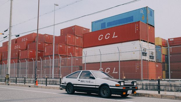 A classic car parked near stacked cargo containers in an industrial area.