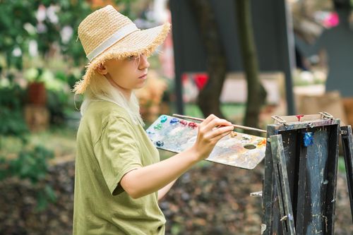 Free A young adult woman artist wearing a straw hat paints outdoors in a summer park setting. Stock Photo