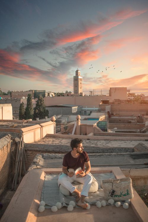 Free Man sitting on a Moroccan rooftop during sunset, with historical minaret in the background. Stock Photo