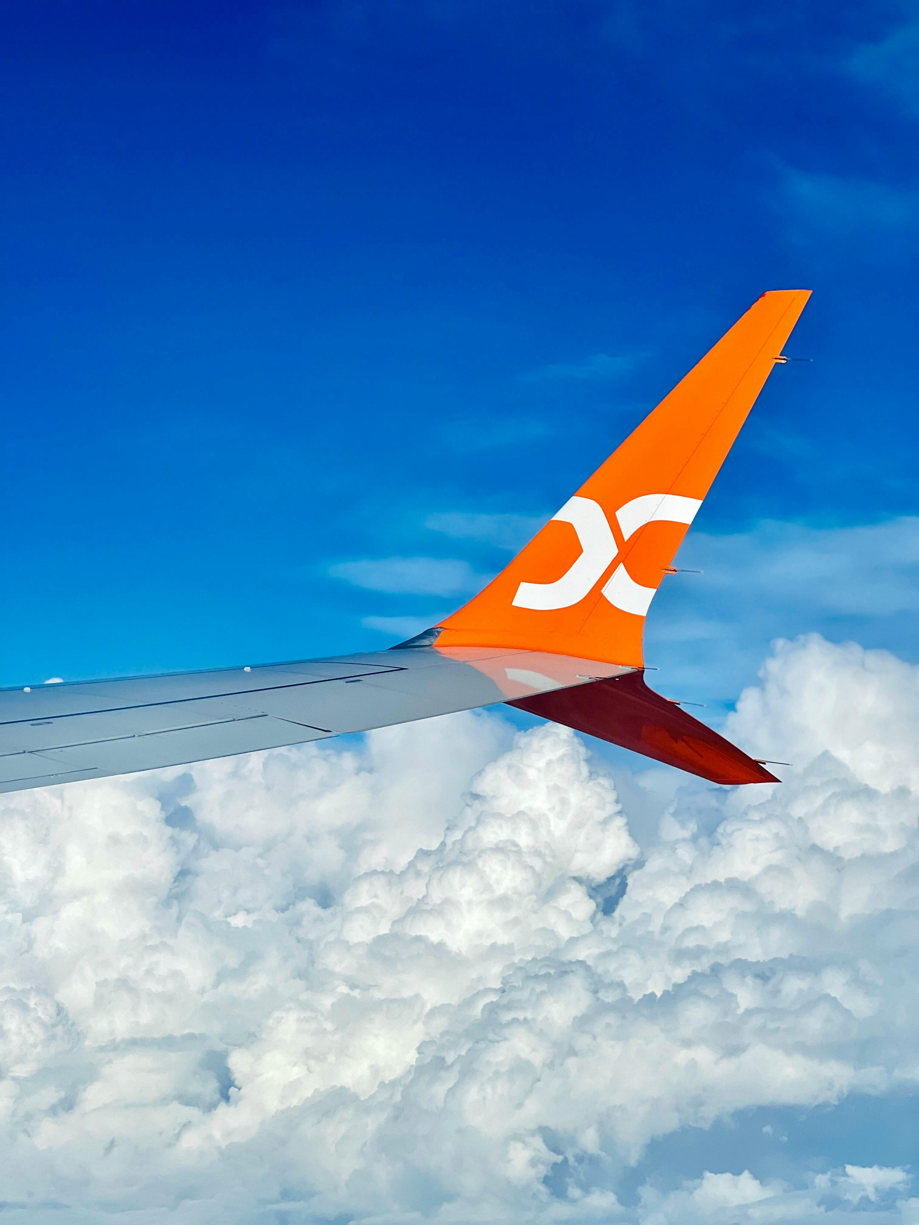 Airplane wing with orange logo flying above fluffy clouds in blue sky.