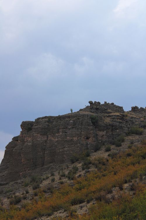 Free A striking rocky cliff rising against a backdrop of a cloudy sky, with autumn foliage below. Stock Photo