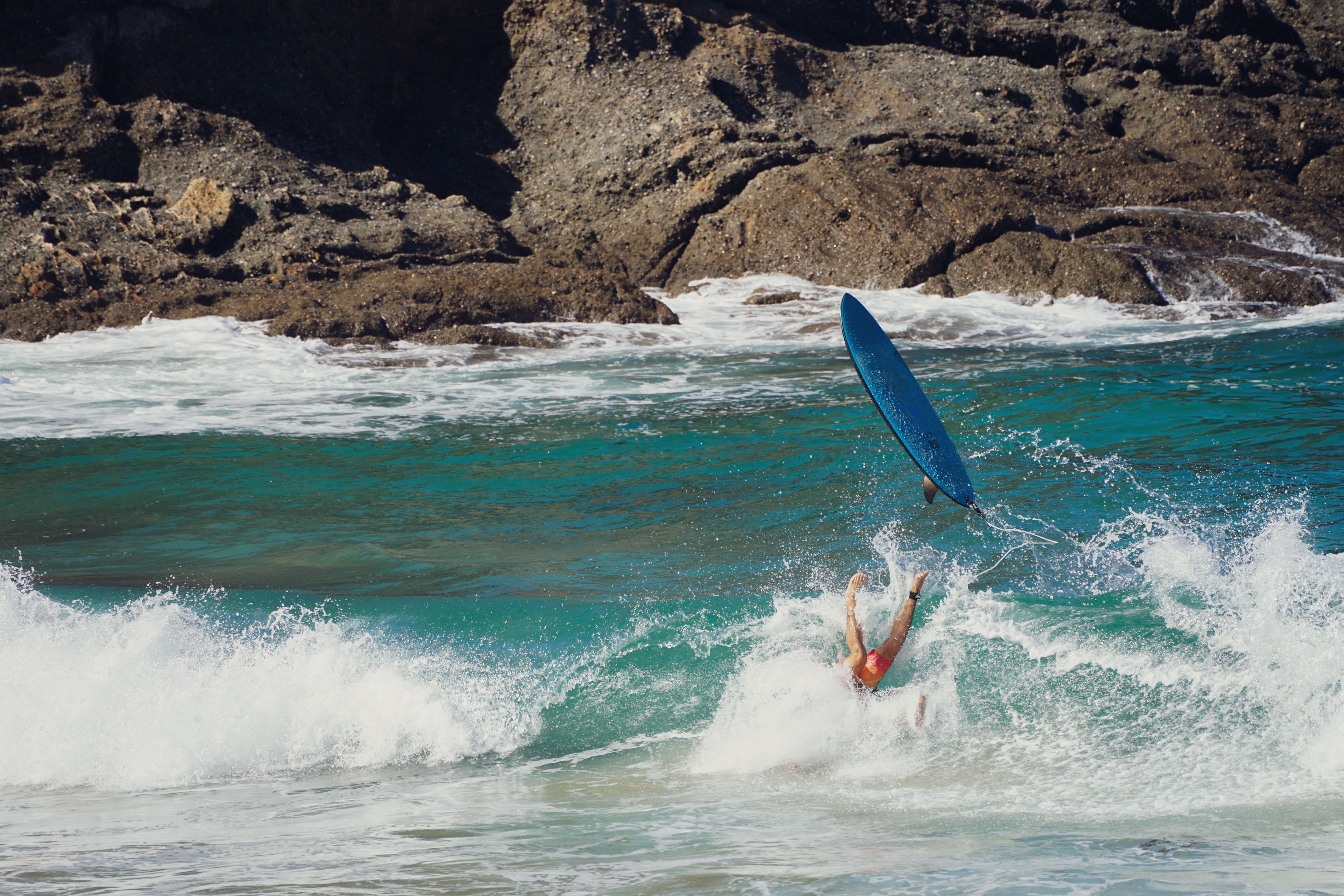 A surfer in vibrant waters takes a fall, surrounded by powerful waves and rugged rocks.