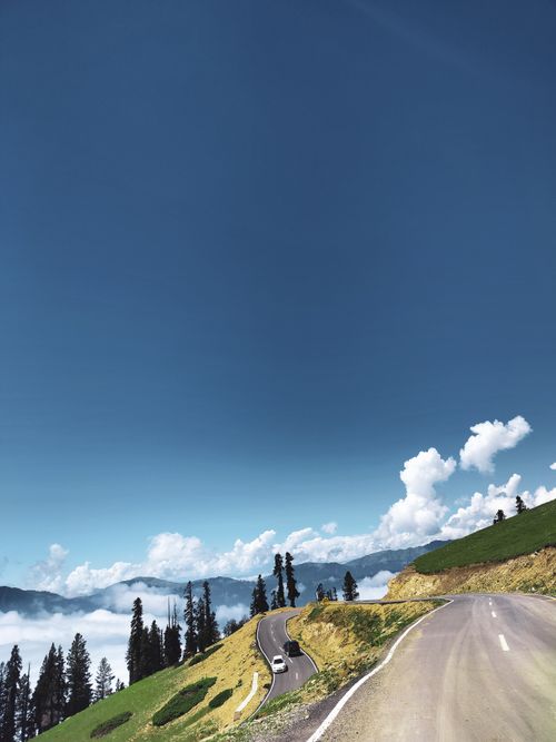 Free A picturesque mountain road meanders through lush greenery under a vibrant blue sky. Stock Photo