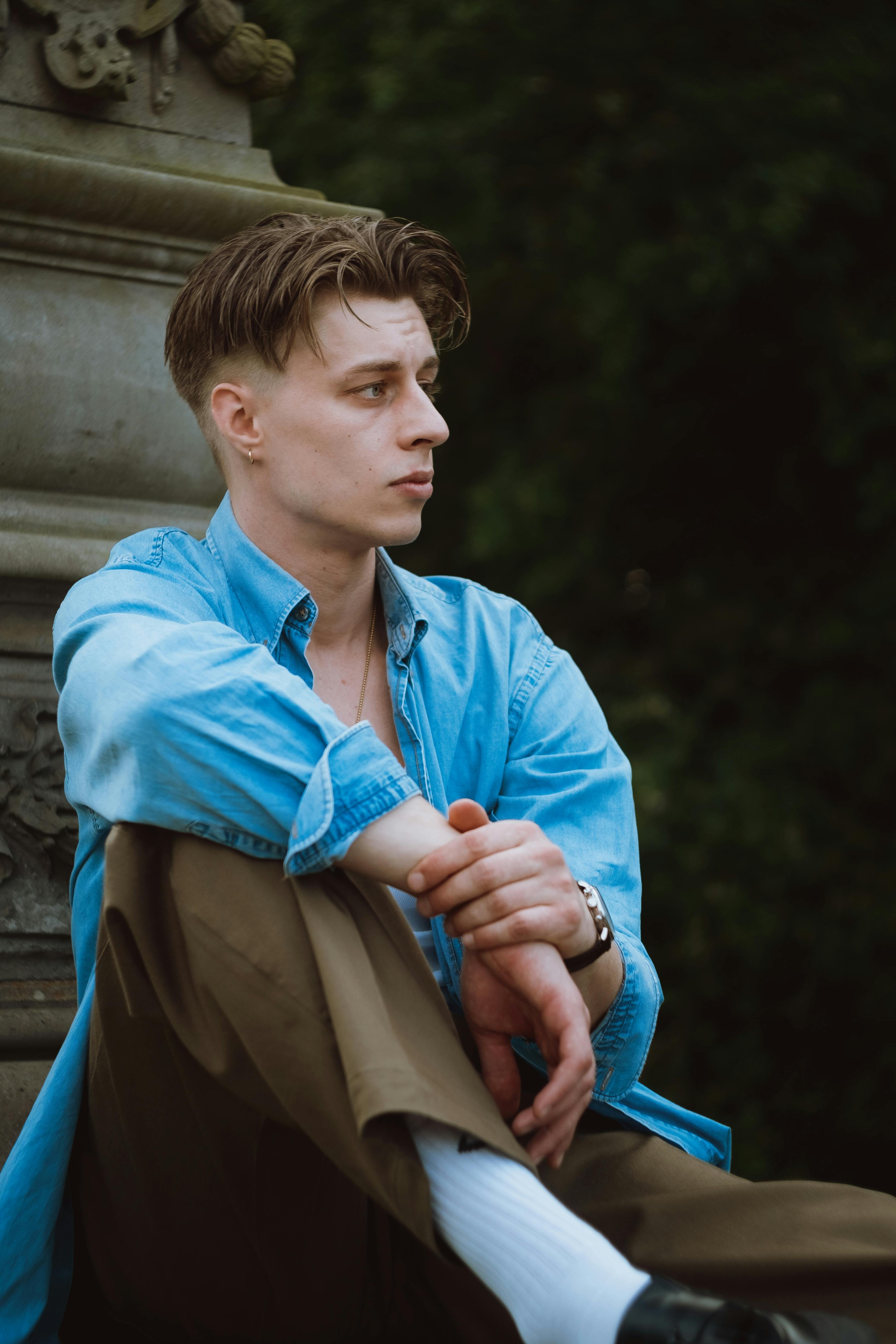 A young man in a blue denim shirt sits pensively outdoors, exuding a vintage fashion vibe.