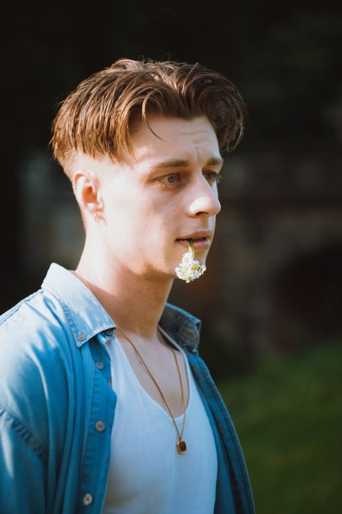 Free A fashionable young man with a flower in his mouth, wearing a denim shirt and necklace outside. Stock Photo