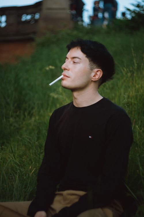 Free Young man sitting in grass, smoking, with a thoughtful expression. Outdoor setting. Stock Photo