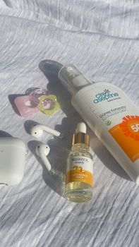 Still life of sunscreen, Vitamin C serum, and earphones on white fabric in sunlight.