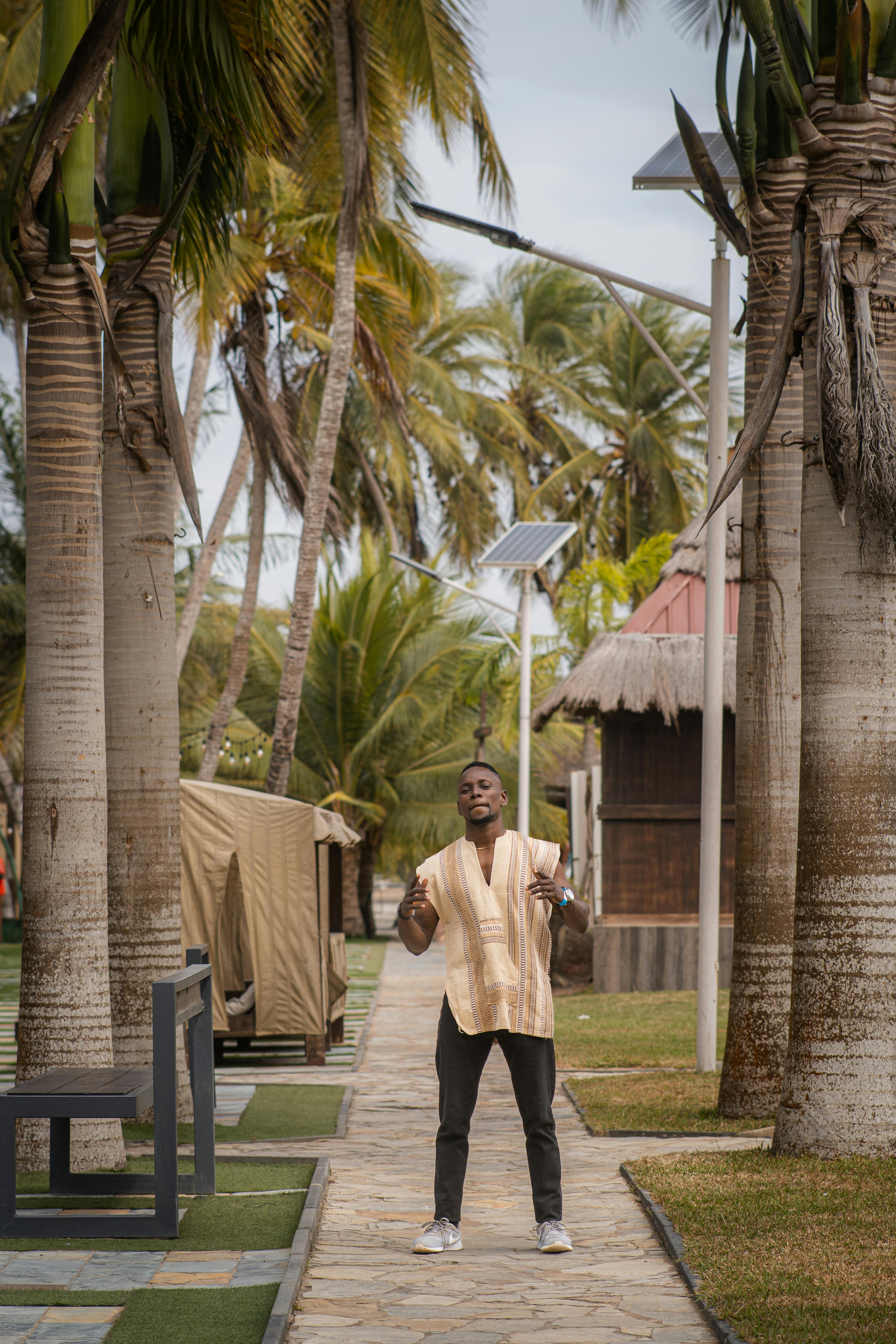 Free Man standing on a pathway in a tropical resort surrounded by palm trees during the day, embracing a leisurely vacation vibe. Stock Photo