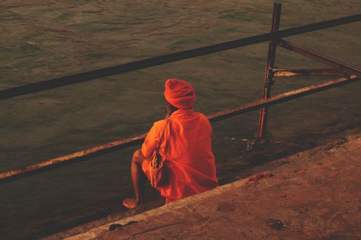A man in traditional attire sits by a riverside, reflecting in solitude.