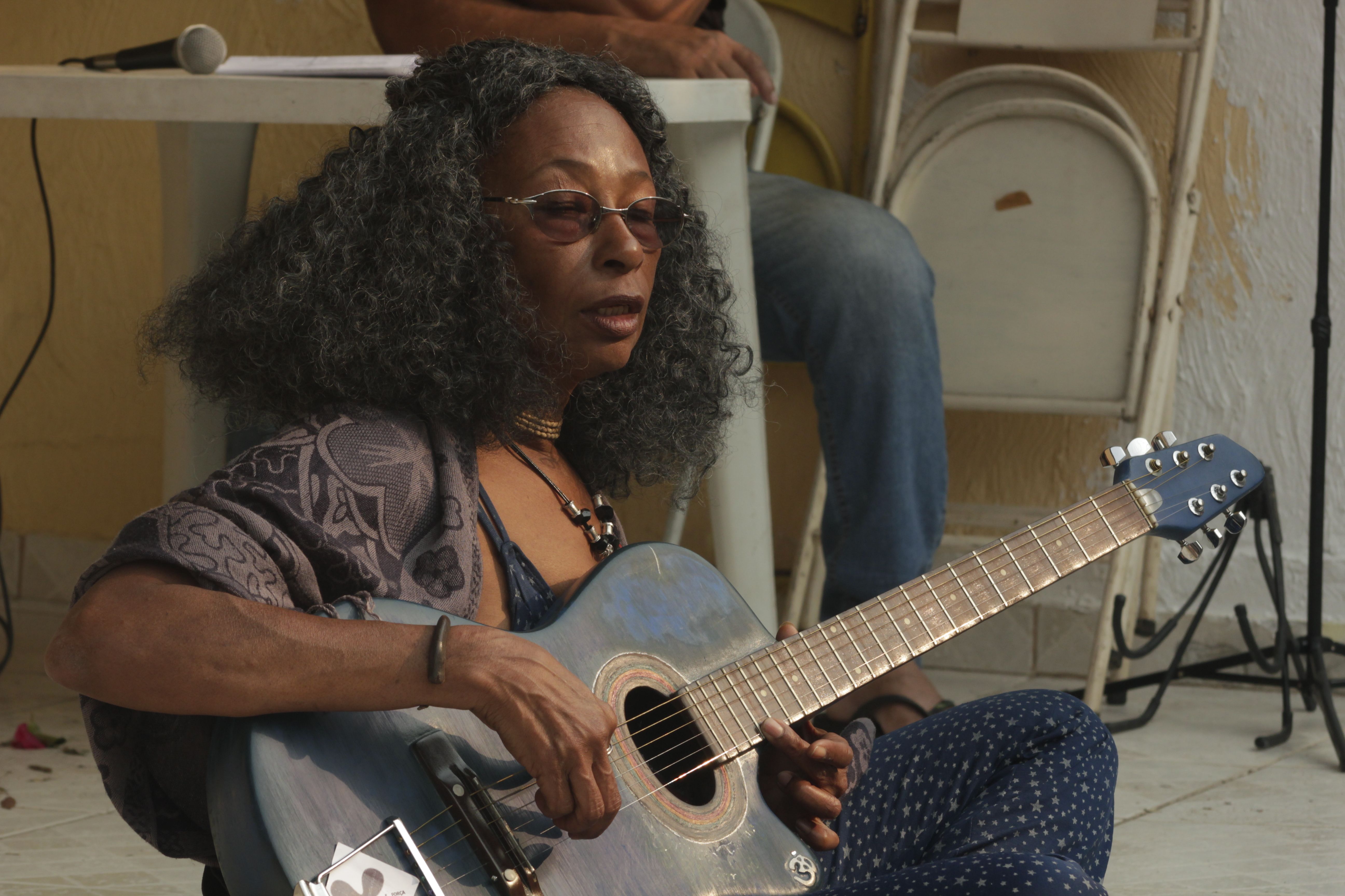 A woman performs outdoors with an acoustic guitar, casual and relaxed.