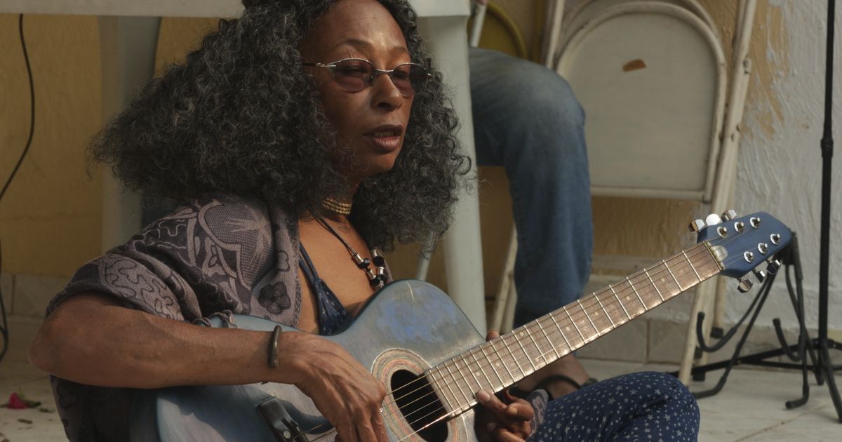 A woman performs outdoors with an acoustic guitar, casual and relaxed.