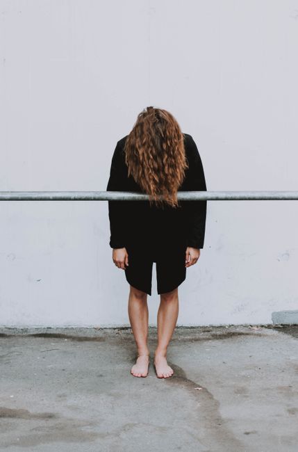 A young woman with long hair stands barefoot and leaning forward against a city wall.
