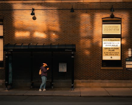 A person waits at a city bus stop by a brick wall under warm lighting. Urban cityscape.
