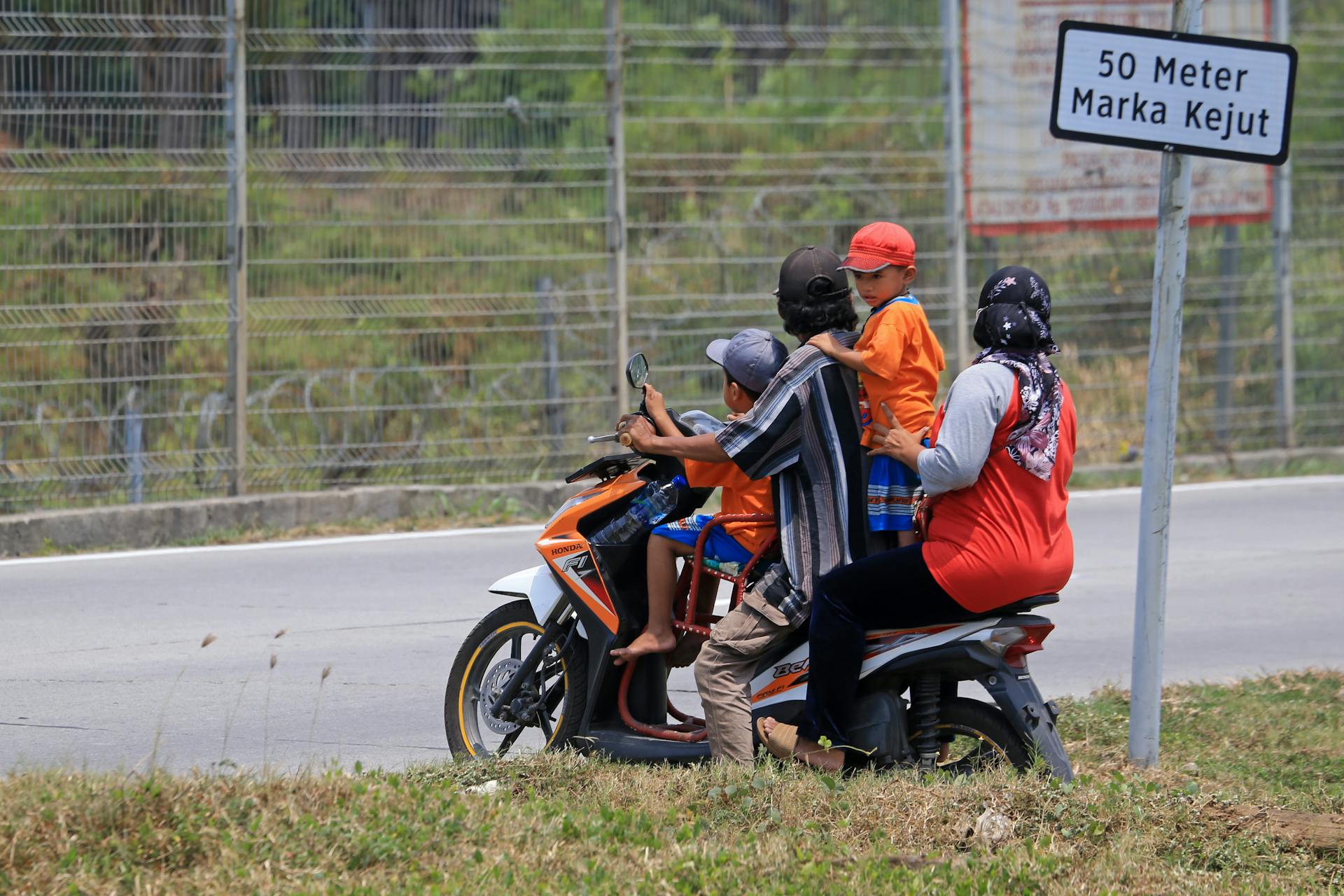 A family travels on a motorcycle along a roadside with a safety sign visible.