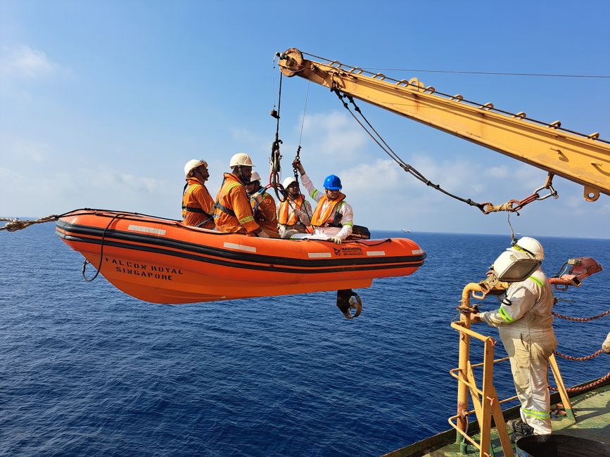 Offshore crew conducting a lifeboat safety drill on a crane over open sea.