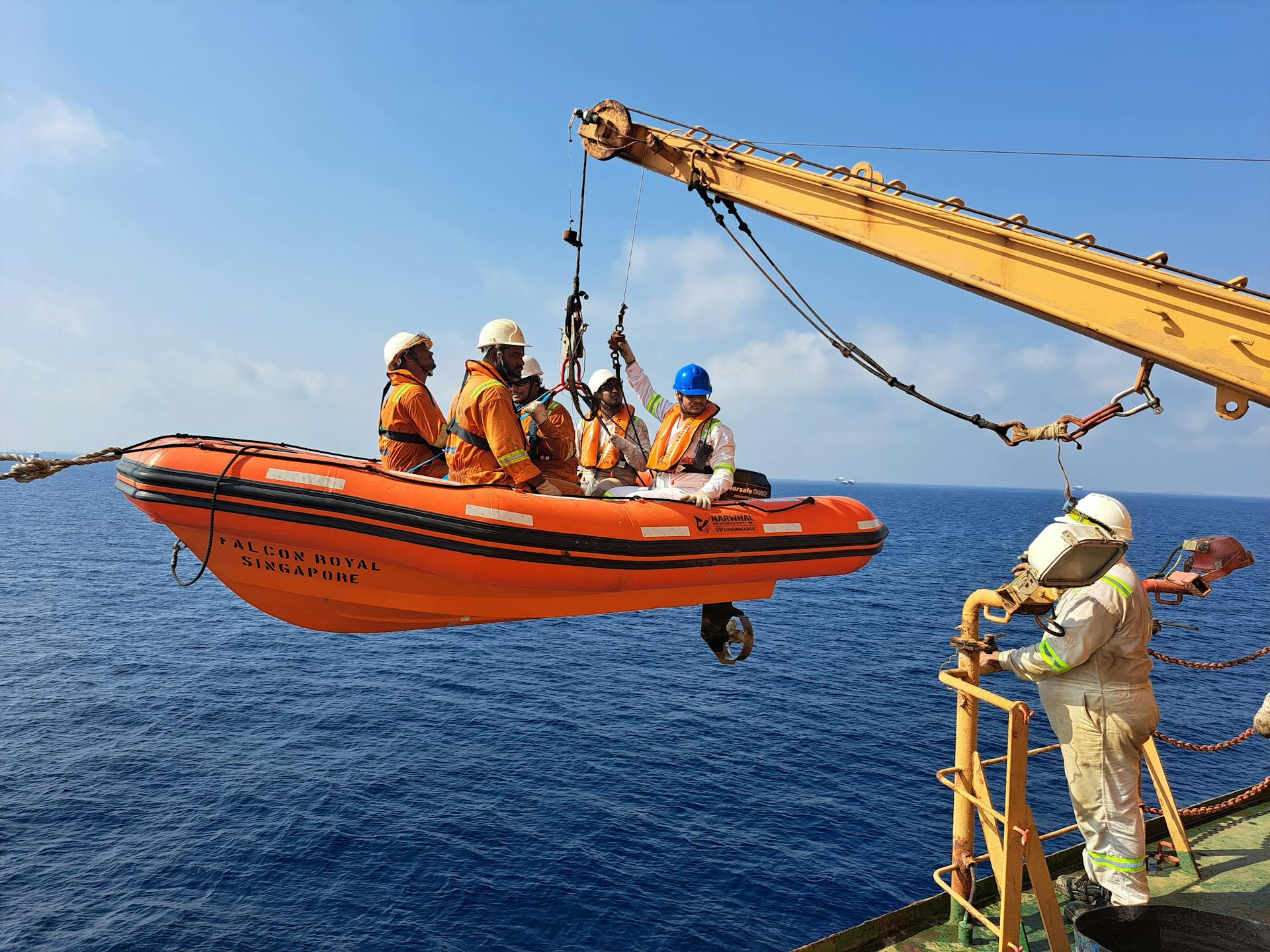 Offshore crew conducting a lifeboat safety drill on a crane over open sea.