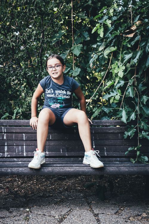 Free Smiling girl sitting on a wooden bench surrounded by lush greenery in a park. Stock Photo