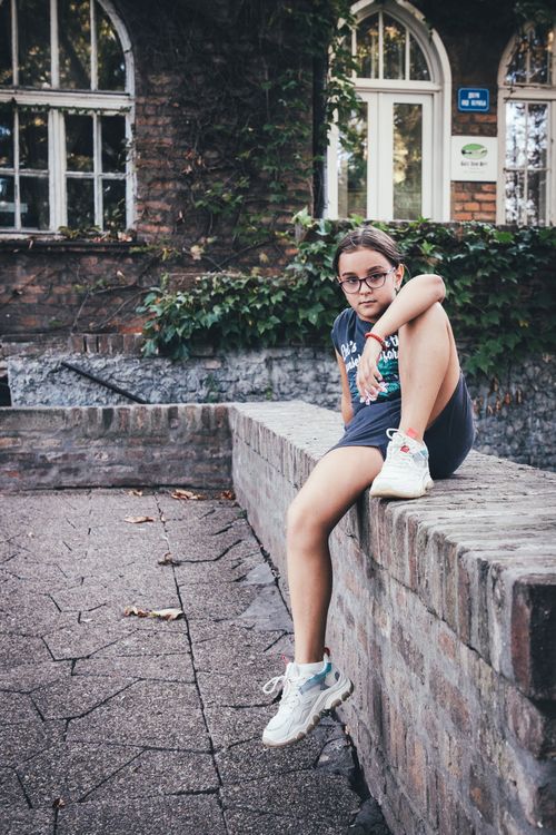 Free A girl casually sits on a brick wall in an urban environment, wearing sneakers and glasses. Stock Photo