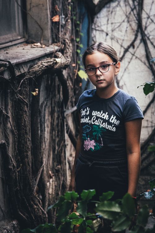 Free Portrait of a girl standing outdoors, surrounded by vines, wearing glasses and a casual t-shirt. Stock Photo