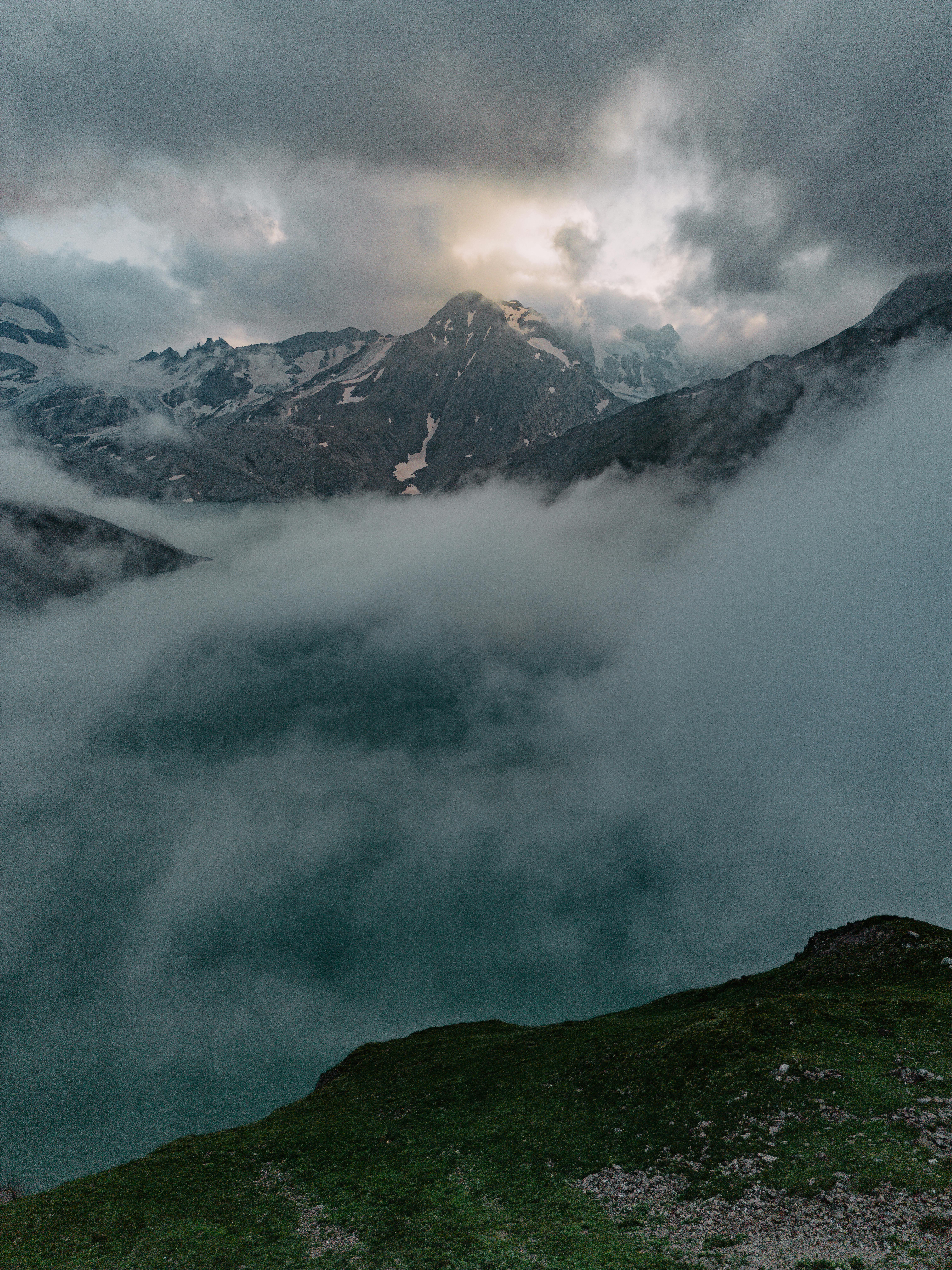 A dramatic mountain landscape with clouds, fog, and snow-capped peaks during daylight.